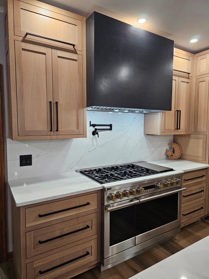 Natural oak shaker cabinetry framing a black wood range hood over a gas range with a marble slab backsplash and pot filler