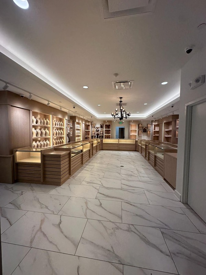 Light oak jewelry showroom with glass display cases, backlit necklace shelving, and marble floors